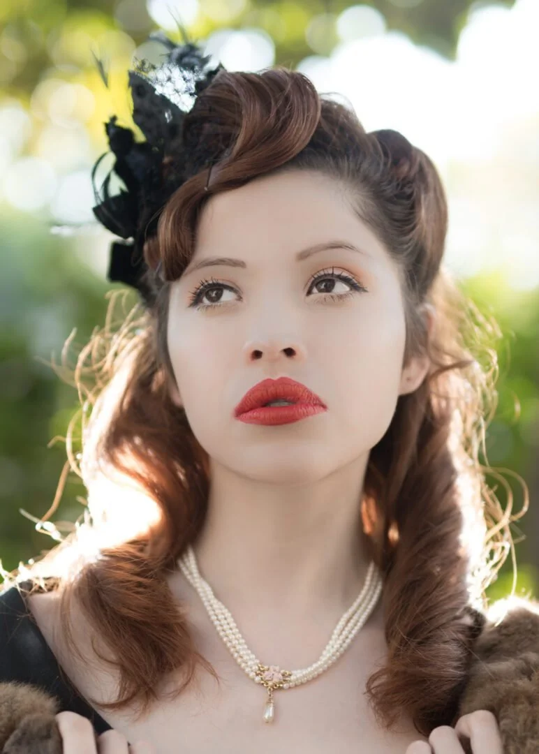 A young woman with wavy, auburn hair wearing a vintage, black floral headpiece, pearl necklace, red lipstick, and a black dress, outdoors with blurred green and yellow foliage in the background.