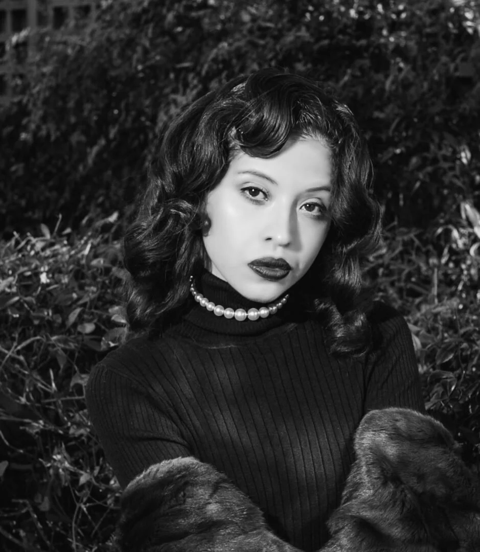 A black and white portrait of a woman with dark, wavy hair, wearing a pearl necklace, a ribbed turtleneck sweater, and a fur coat, standing outdoors with foliage in the background.