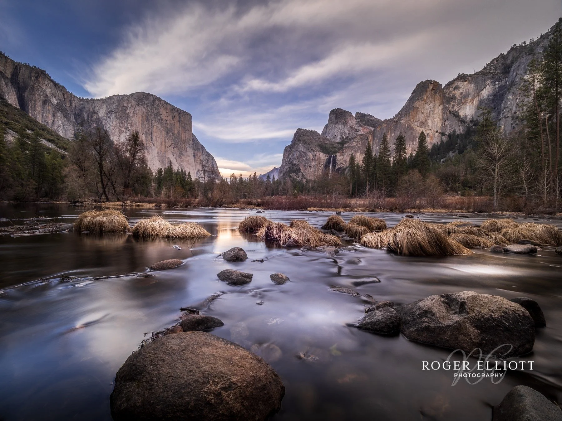 A scenic landscape of Yosemite National Park featuring a river with rocks and patches of grass, surrounded by tall trees and rugged cliffs under a partly cloudy sky.