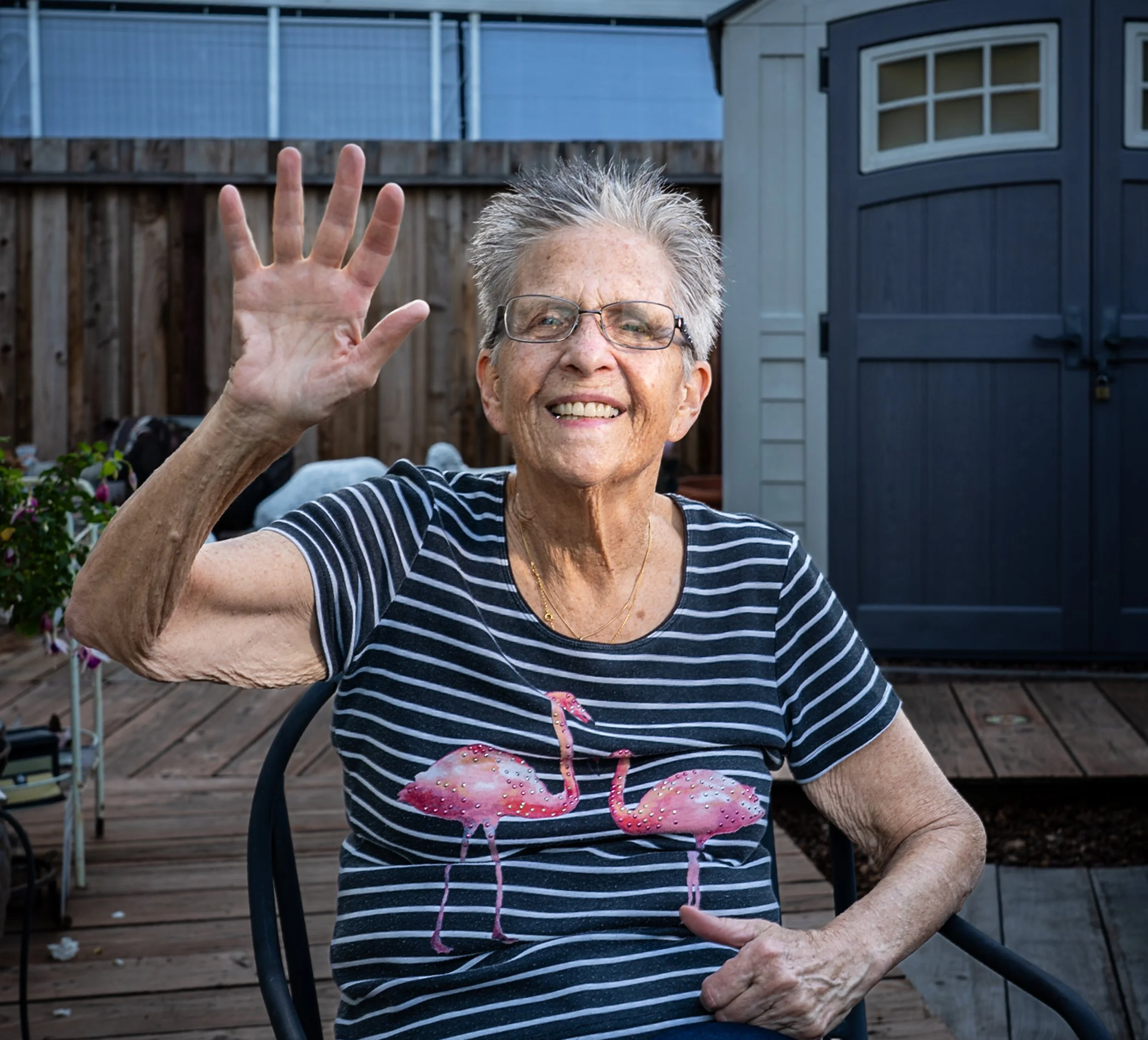 A smiling elderly woman with short gray hair and glasses, wearing a striped T-shirt with flamingos, waving outside on a wooden deck.