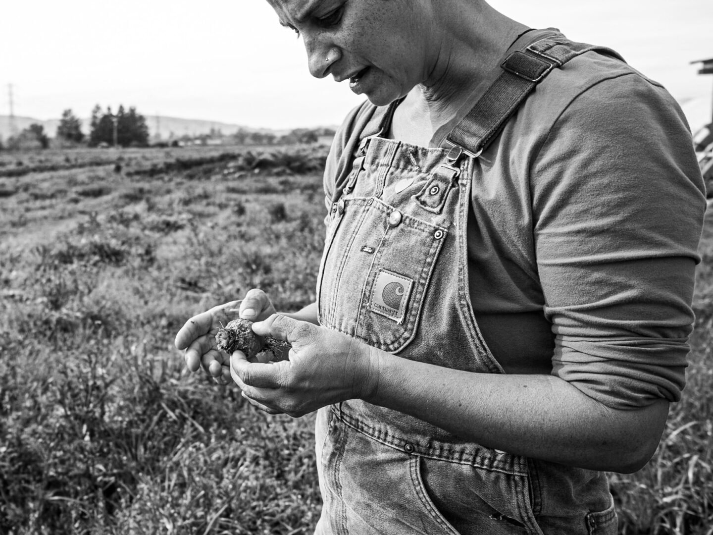 A person wearing a Carhartt denim overall and a long-sleeve shirt holds a small plant or seedling in their hands while standing outdoors in a field, with trees and power lines visible in the background.