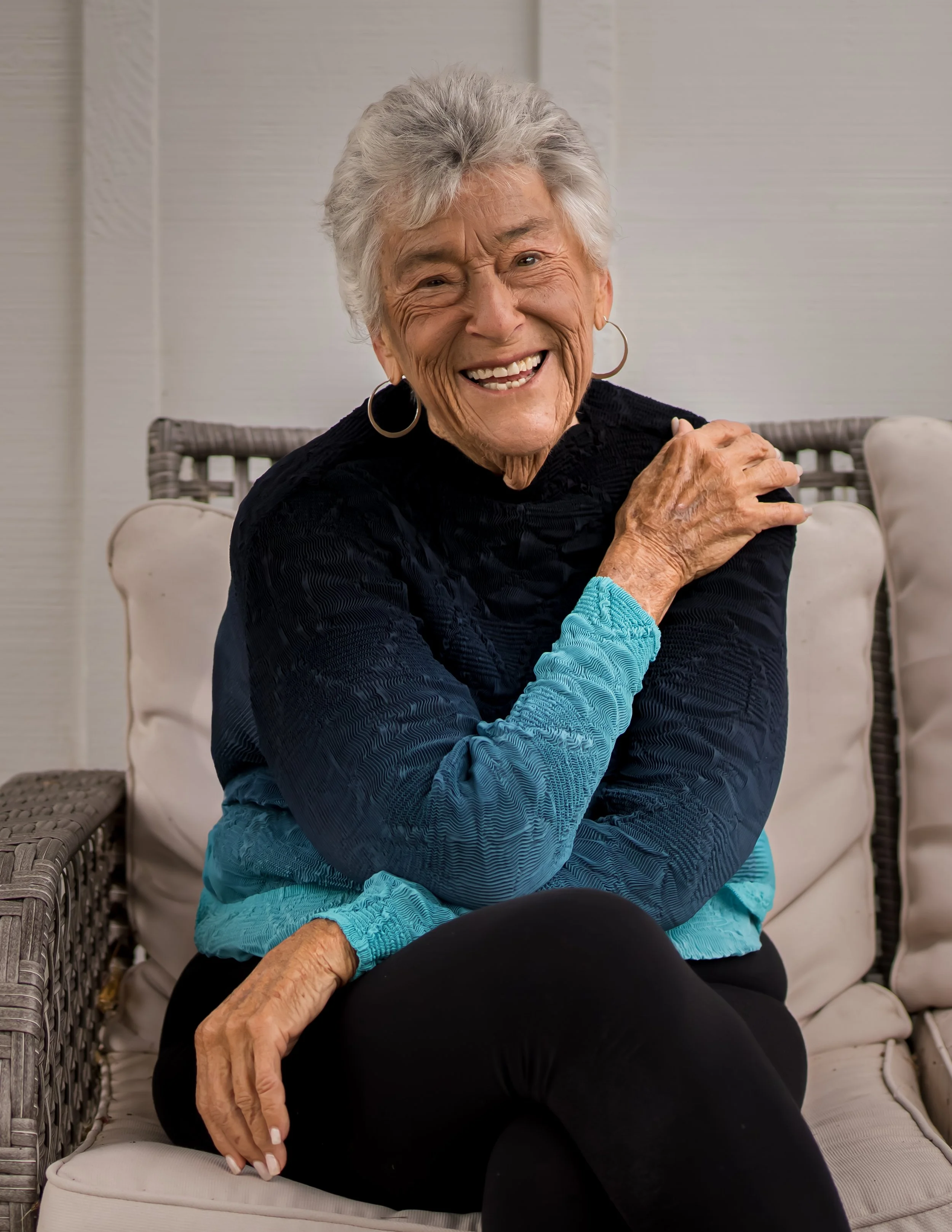 An elderly woman with gray hair, wearing hoop earrings and a black and blue sweater, smiling while sitting on outdoor furniture.