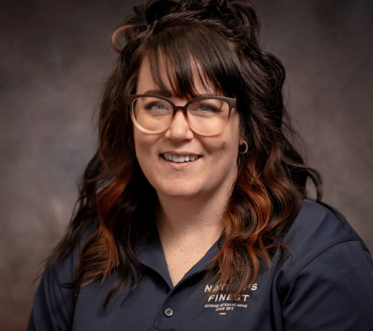 A woman with long dark hair, wearing glasses and a navy blue shirt, smiling against a dark, neutral background.