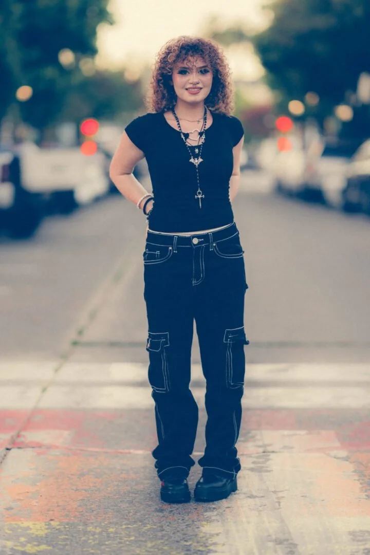A young woman with curly hair standing on a crosswalk in an urban area, smiling at the camera.
