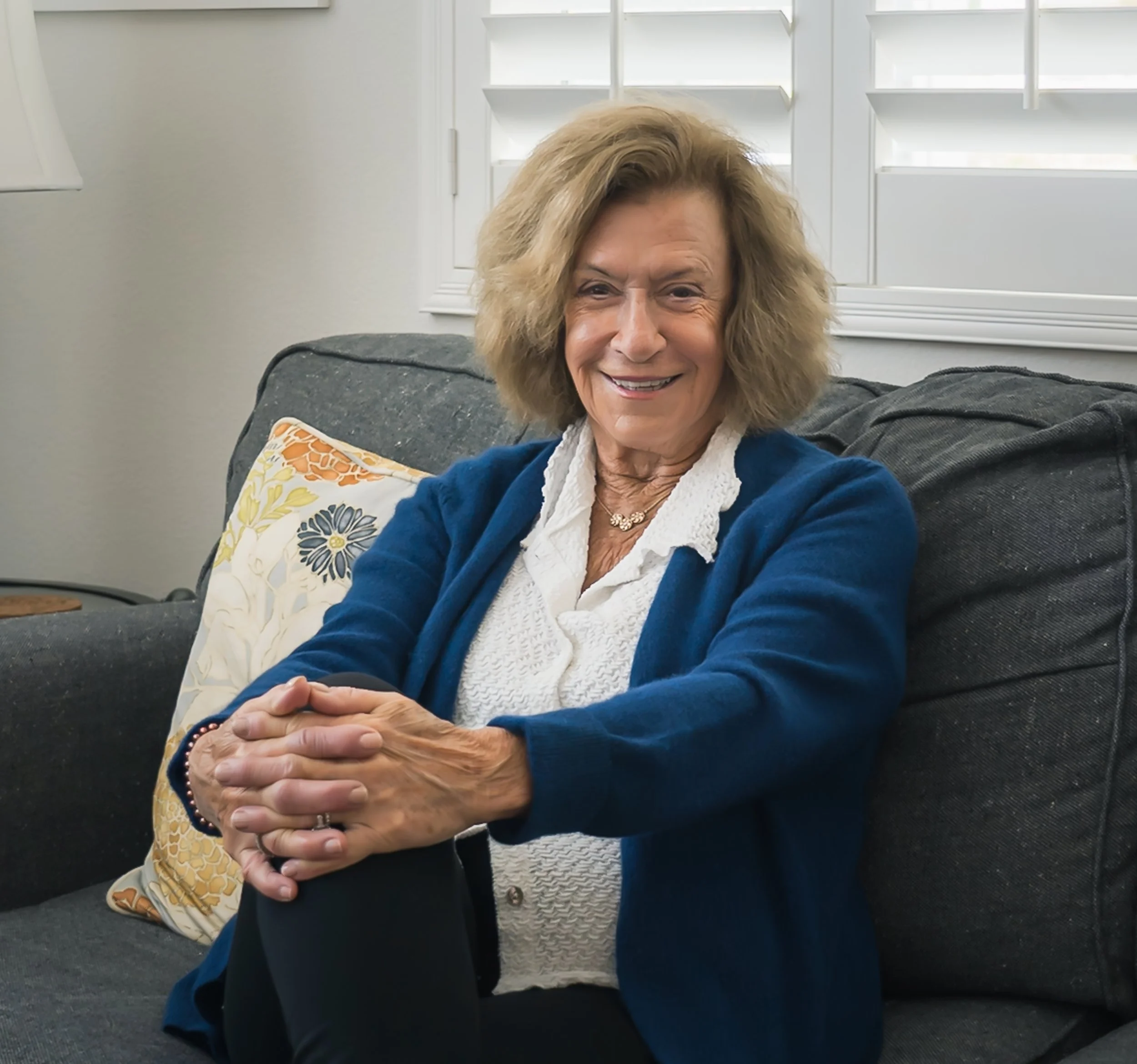 An elderly woman with curly blonde hair, wearing a blue cardigan over a white blouse, sitting on a dark gray couch with a decorative pillow in a well-lit room with white walls and window shutters, smiling at the camera.