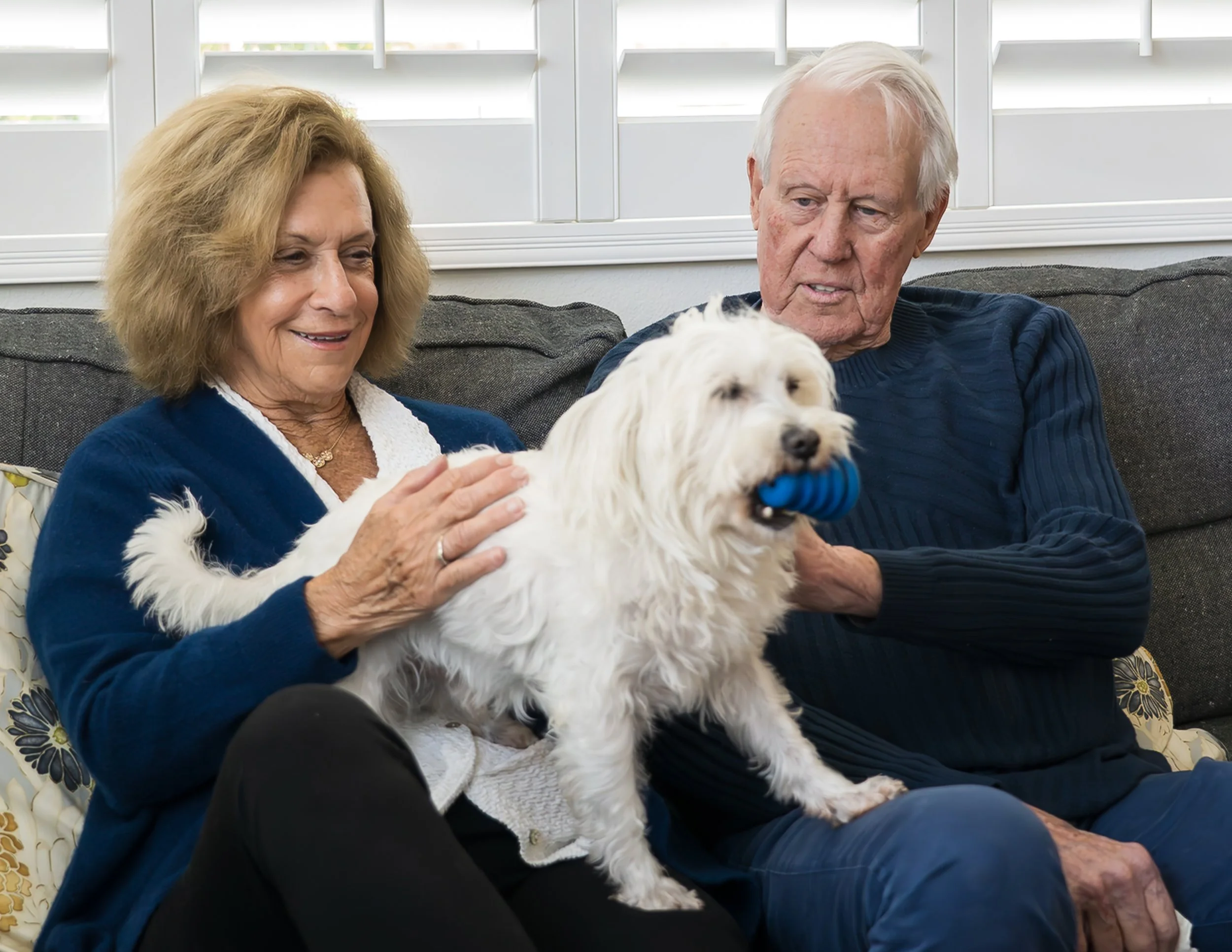 An elderly woman and man sitting on a sofa with a white dog holding a blue chew toy in its mouth. The woman is petting the dog, and they are in a room with white window blinds.