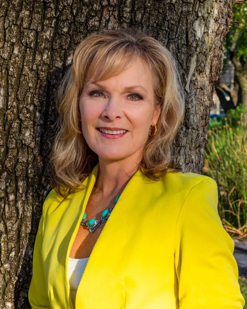 A woman with blonde hair smiling, standing outdoors against a tree trunk, wearing a yellow blazer and turquoise jewelry.