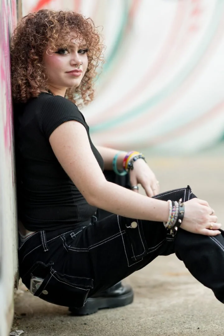A young woman with curly red hair, wearing a black t-shirt and black jeans, sits against a colorful wall looking at the camera. She has bracelets on her wrists and a septum piercing.