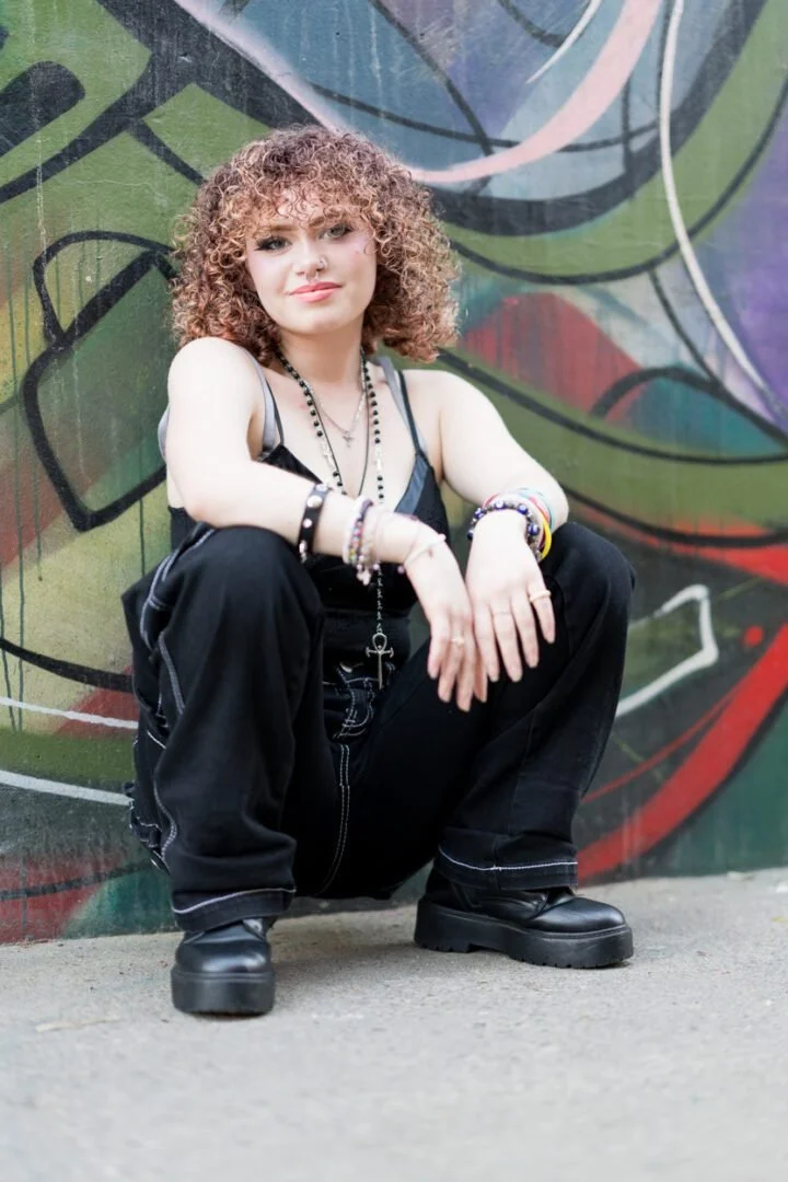 A young woman with curly hair squatting in front of a colorful graffiti wall, wearing black clothing and boots, with multiple bracelets and necklaces.