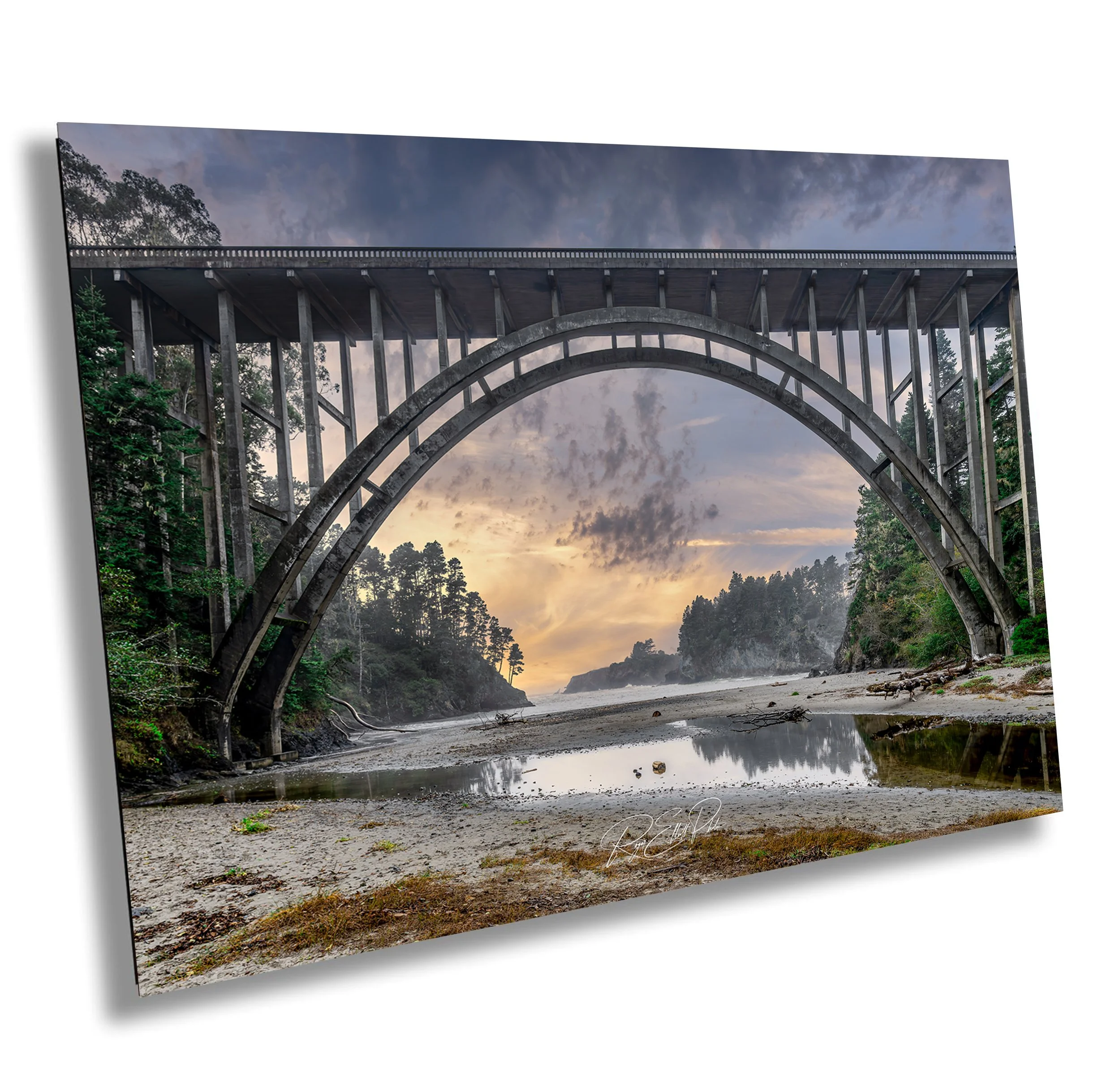 A large concrete arch bridge spans over a rugged, sandy riverbed with small pools of water and driftwood. Tall green trees surround the scene, and the sky shows a colorful sunset with clouds.