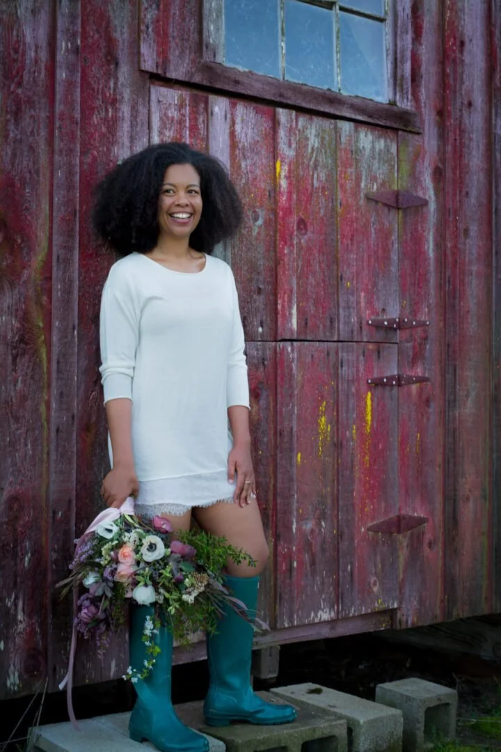 A woman with curly black hair wearing a white long-sleeve shirt and teal rain boots, holding a bouquet of flowers, standing in front of a weathered, red wooden barn with a slightly open door.