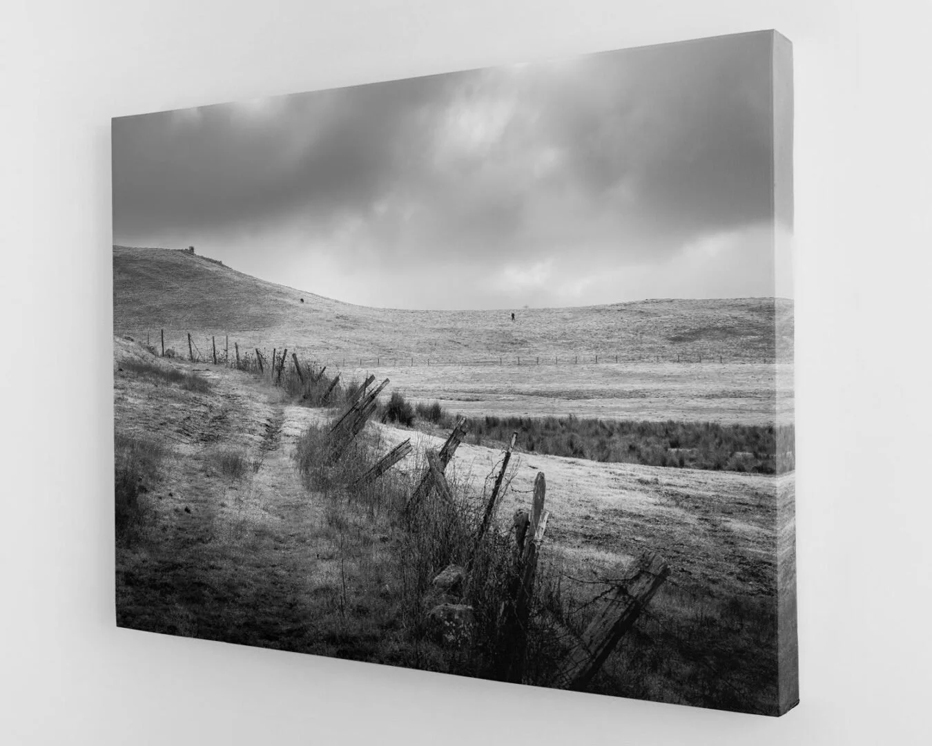 Black and white landscape photograph of a hilly countryside with a fence running along a dirt path, dark clouds overhead, and a solitary figure in the distance.