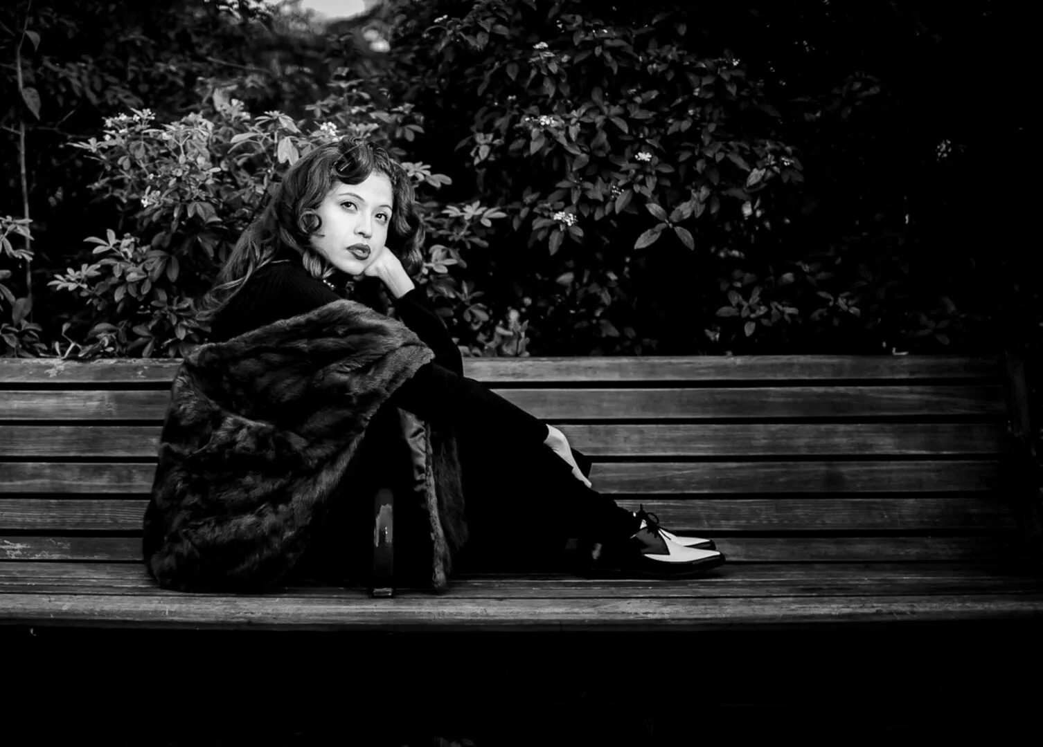 A woman sitting on a wooden park bench, wearing vintage attire with a fur stole, surrounded by bushes with small flowers, in black and white photography.