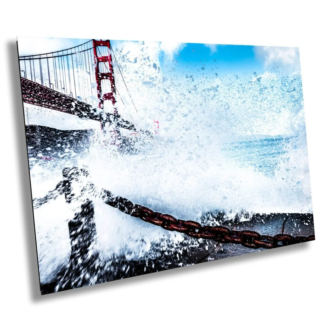 The Golden Gate Bridge with seawater splashing and a chain in the foreground, under a partly cloudy sky.