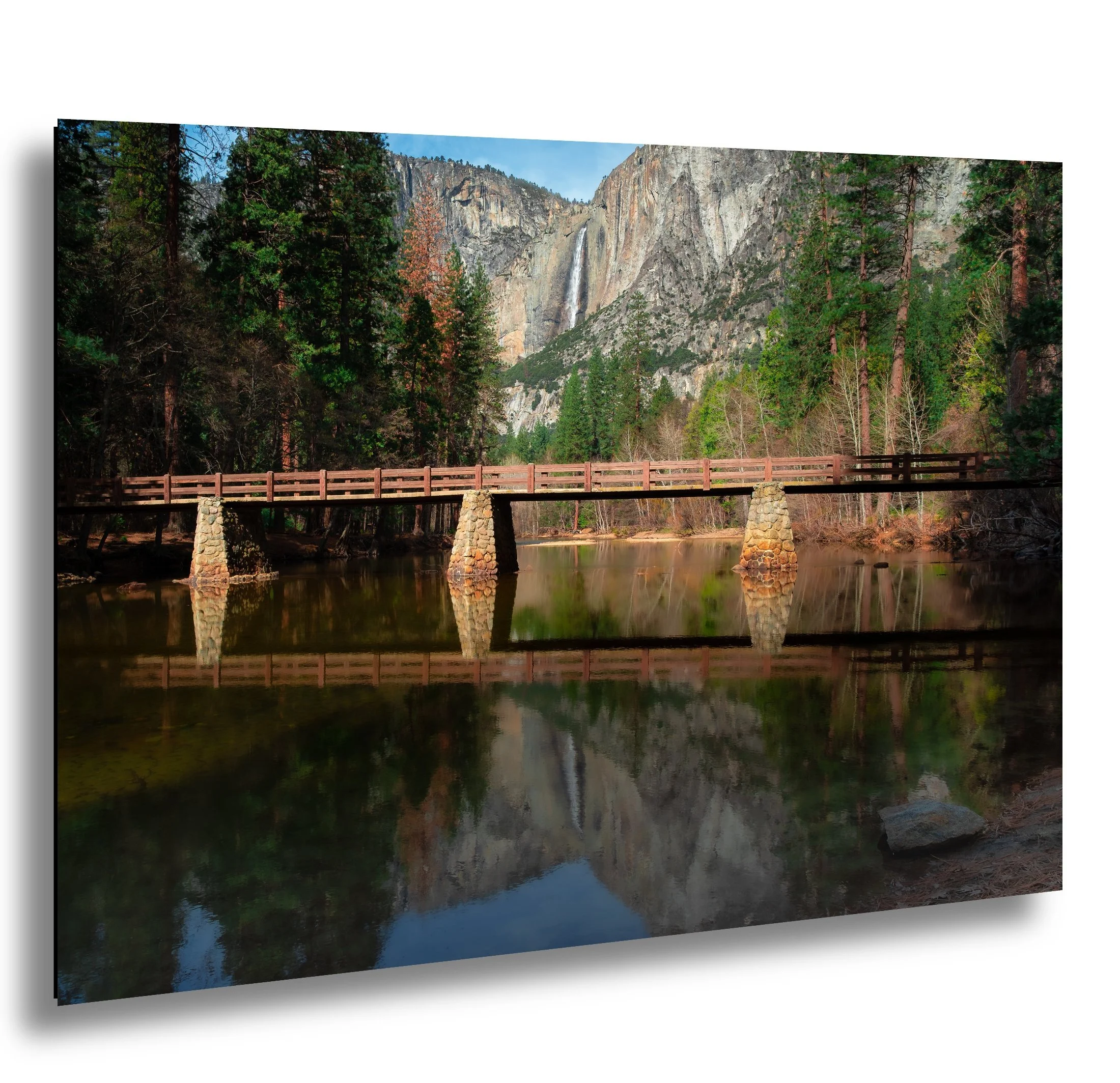 A scenic view of a wooden bridge over a calm river with mountains, trees, and a waterfall in the background.