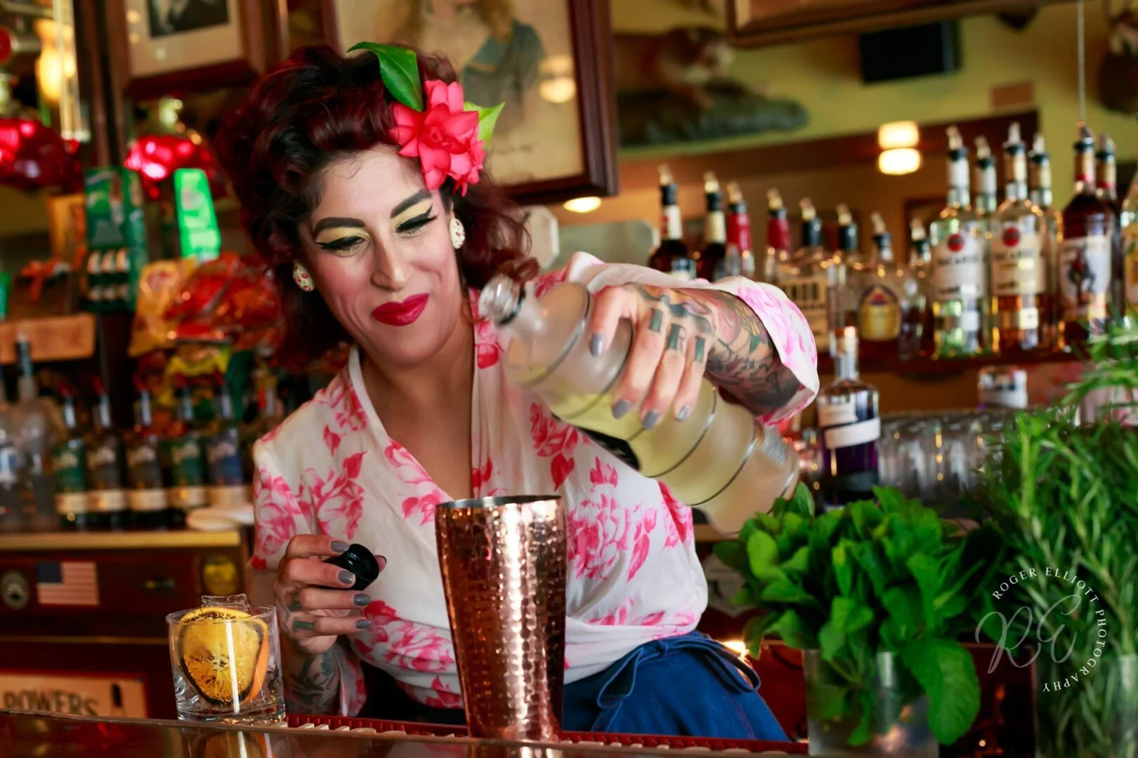 A woman with tattoos and with makeup and a floral hair accessory is pouring a clear beverage with lemon into a copper cup at a bar.