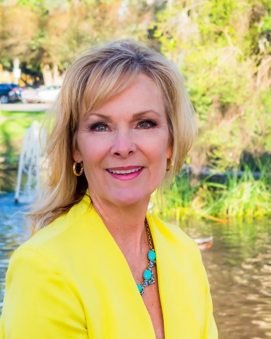 A smiling woman with blonde hair wearing a yellow blazer and turquoise jewelry standing outdoors near a pond with trees and a fountain in the background.