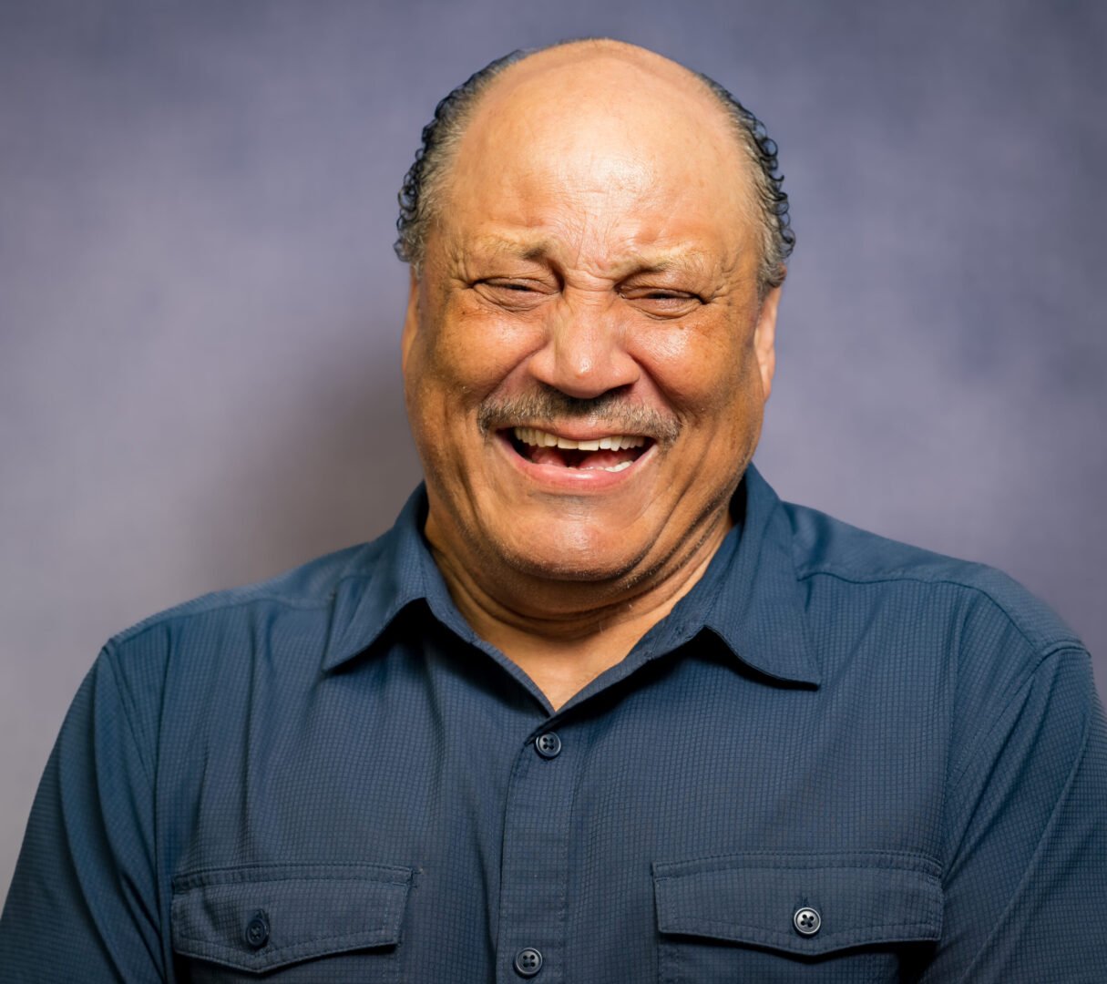 An elderly man with a bald head, gray hair on the sides, and a mustache is smiling while laughing. He is wearing a dark blue button-up shirt and stands against a plain gray background.