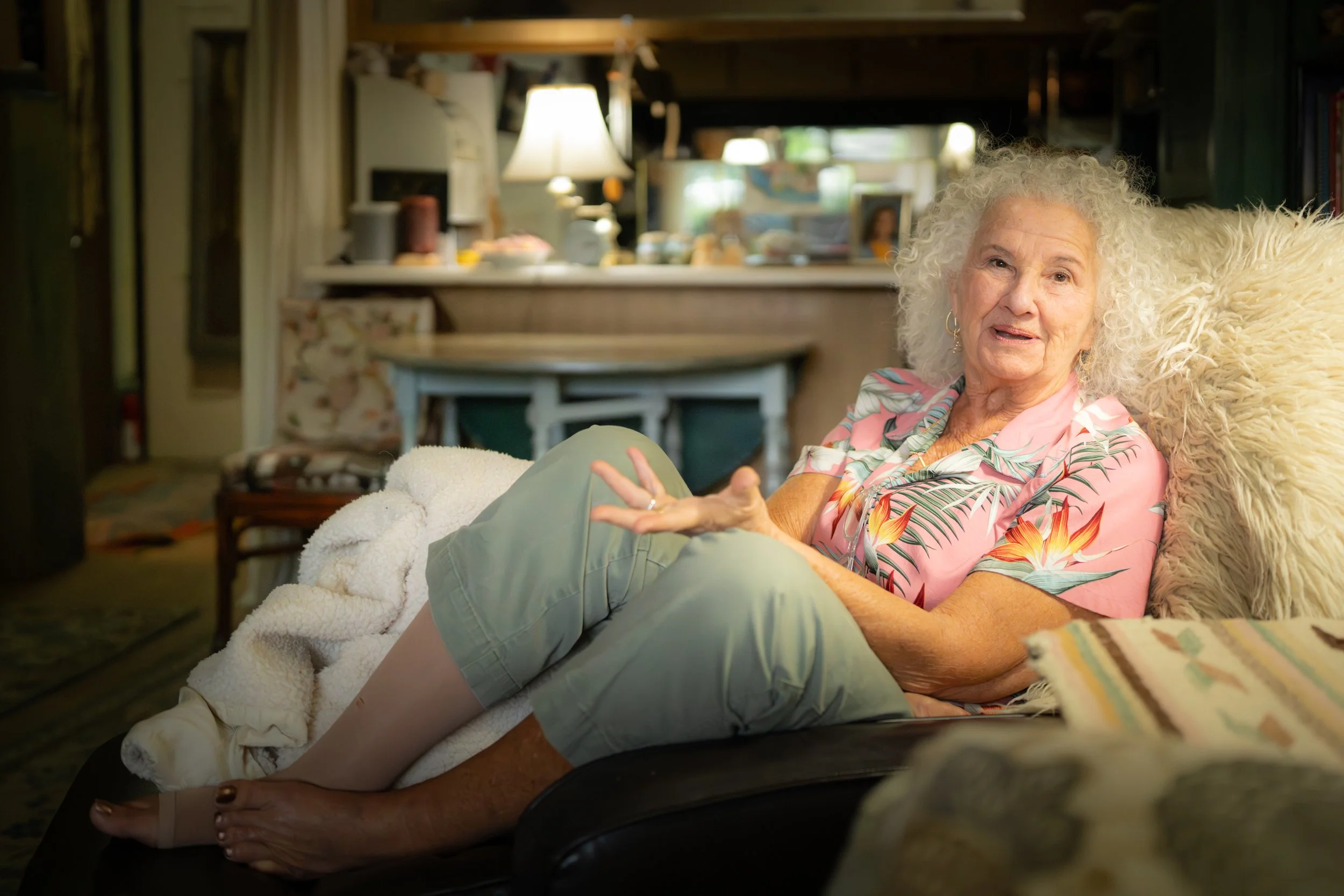 An elderly woman with curly gray hair sitting on a couch in a cozy, well-lit living room, wearing a pink floral shirt and green capris, gesturing with her hand and looking at the camera.