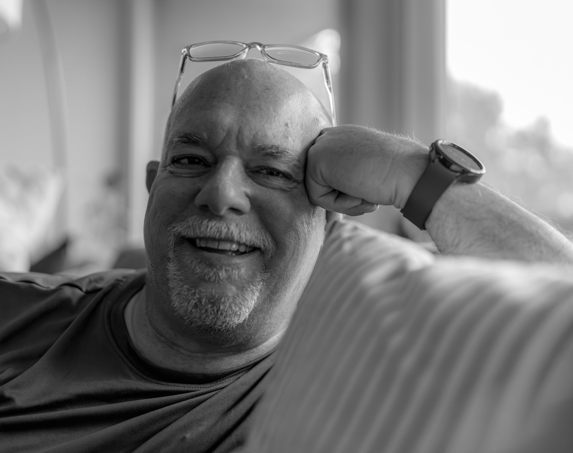 A smiling, bald man with a goatee, wearing eyeglasses on his head, a watch, and a casual shirt, resting his head on his hand while sitting on a striped couch.
