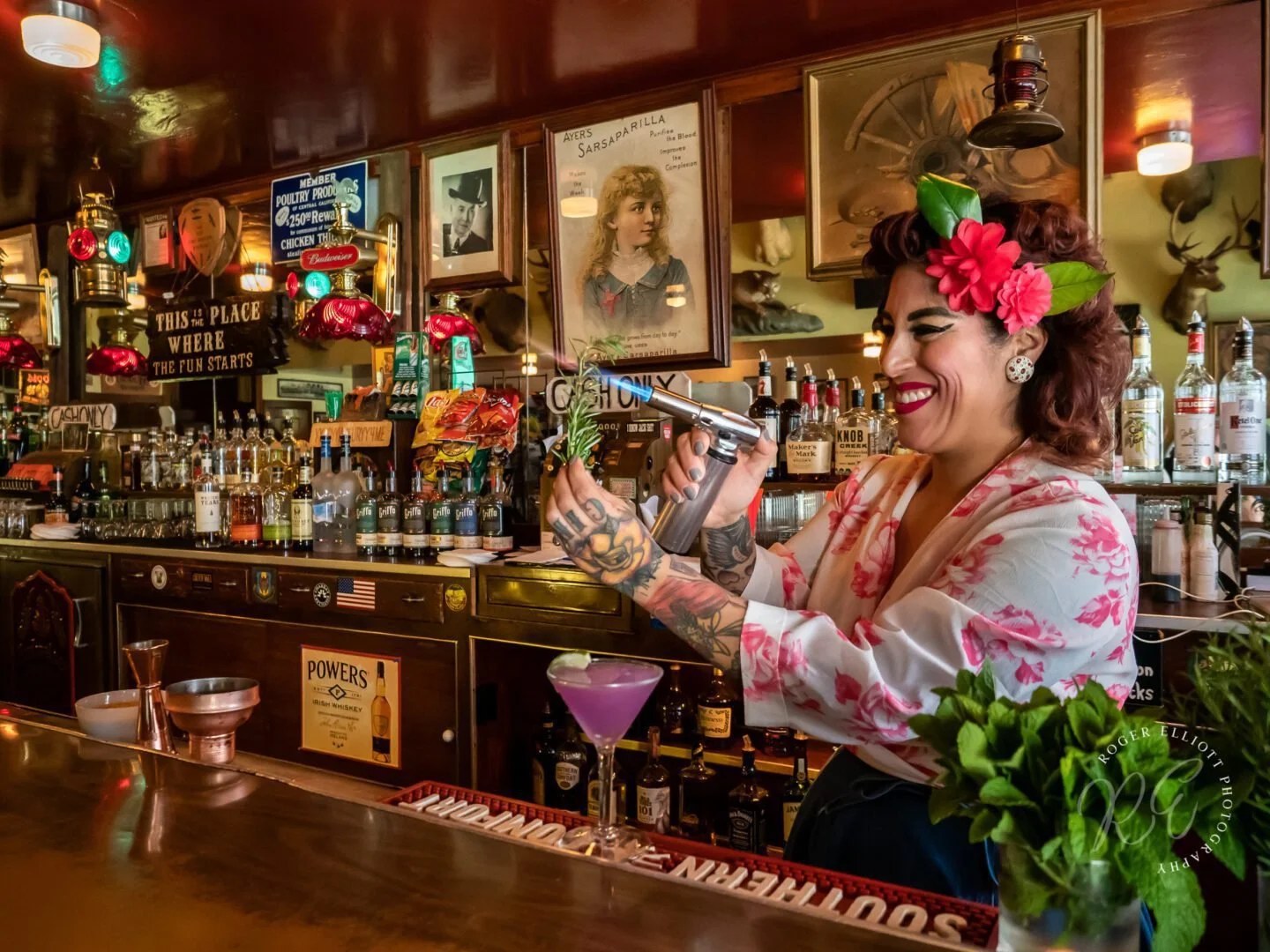 A smiling woman with tattoos, wearing floral earrings, a pink floral blouse, and a flower headpiece, prepares a cocktail behind a bar counter in a vintage-themed bar with liquor bottles and framed pictures.