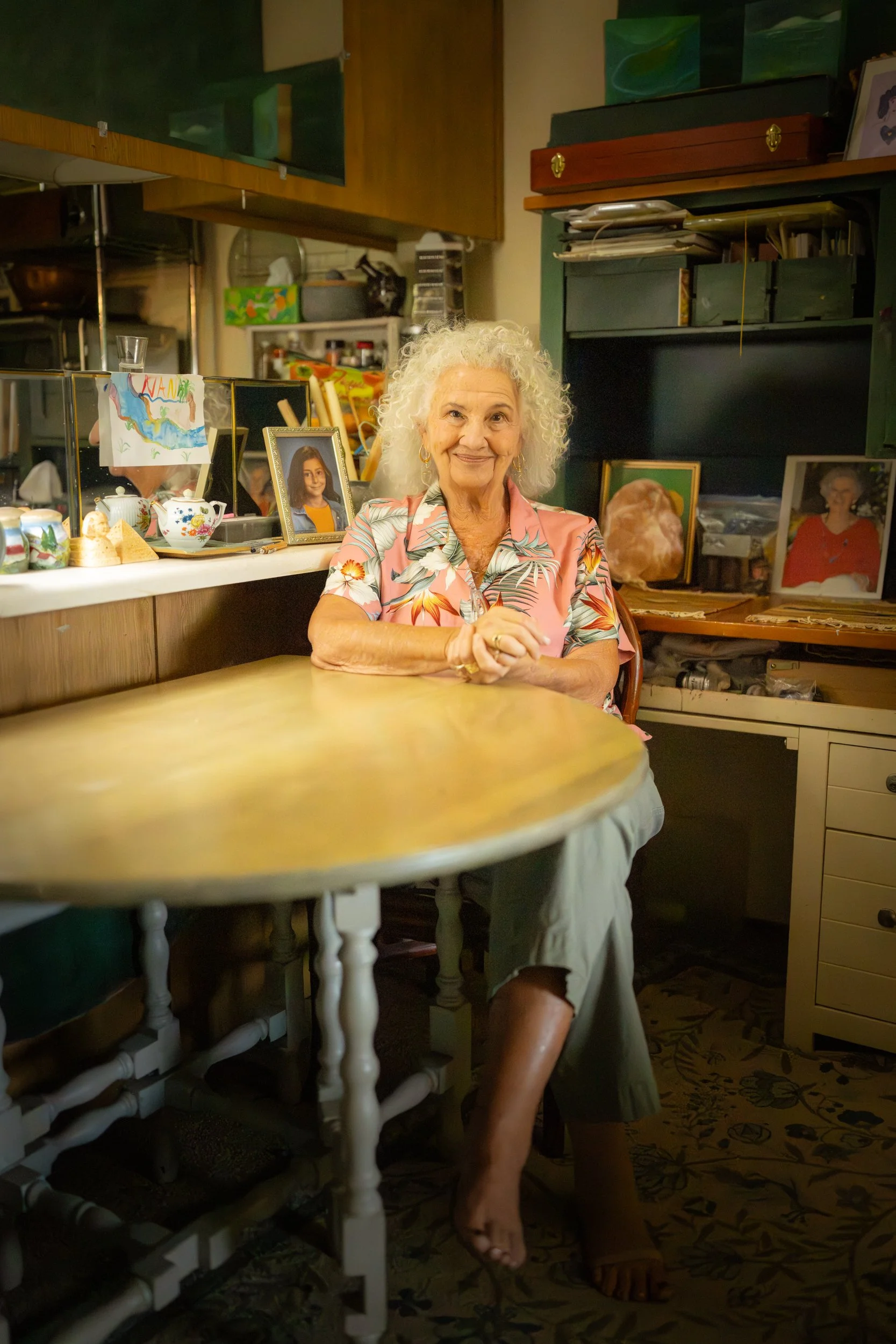 An elderly woman with curly gray hair sitting at a wooden table in a cozy room decorated with family photos and artwork.
