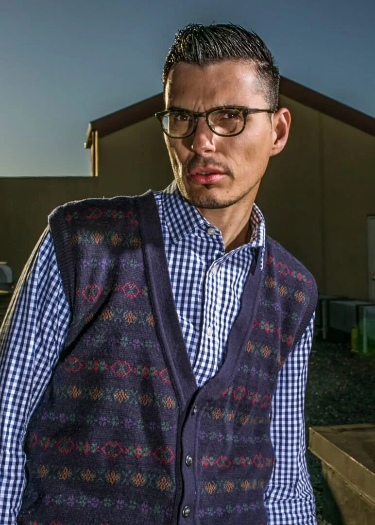 A man with glasses, wearing a blue and white checkered shirt and a dark sweater vest with patterns, standing outdoors in the evening with a building and a dark sky in the background.