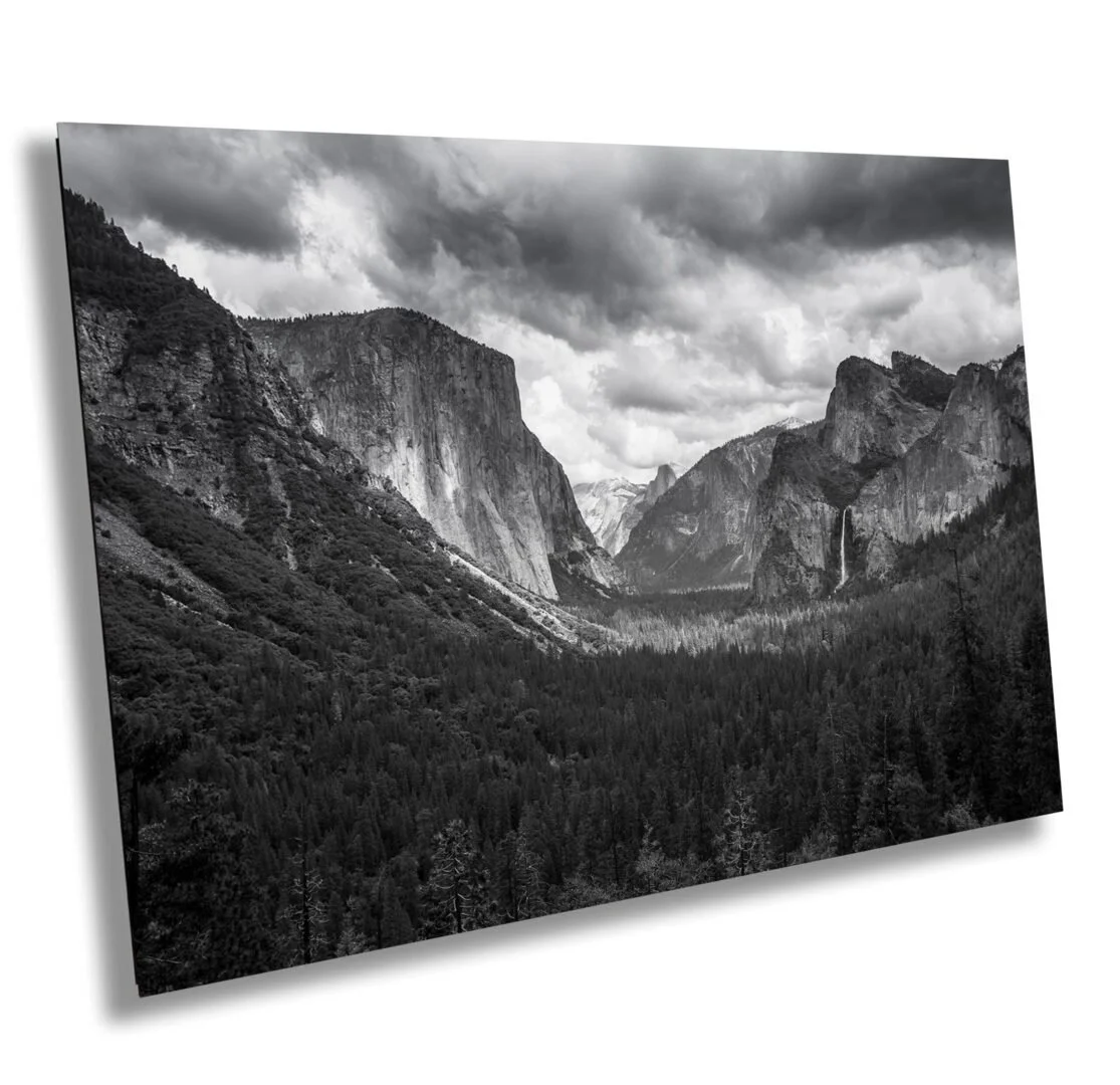Black and white photograph of a mountain landscape with steep cliffs, forested valleys, and a cloudy sky.
