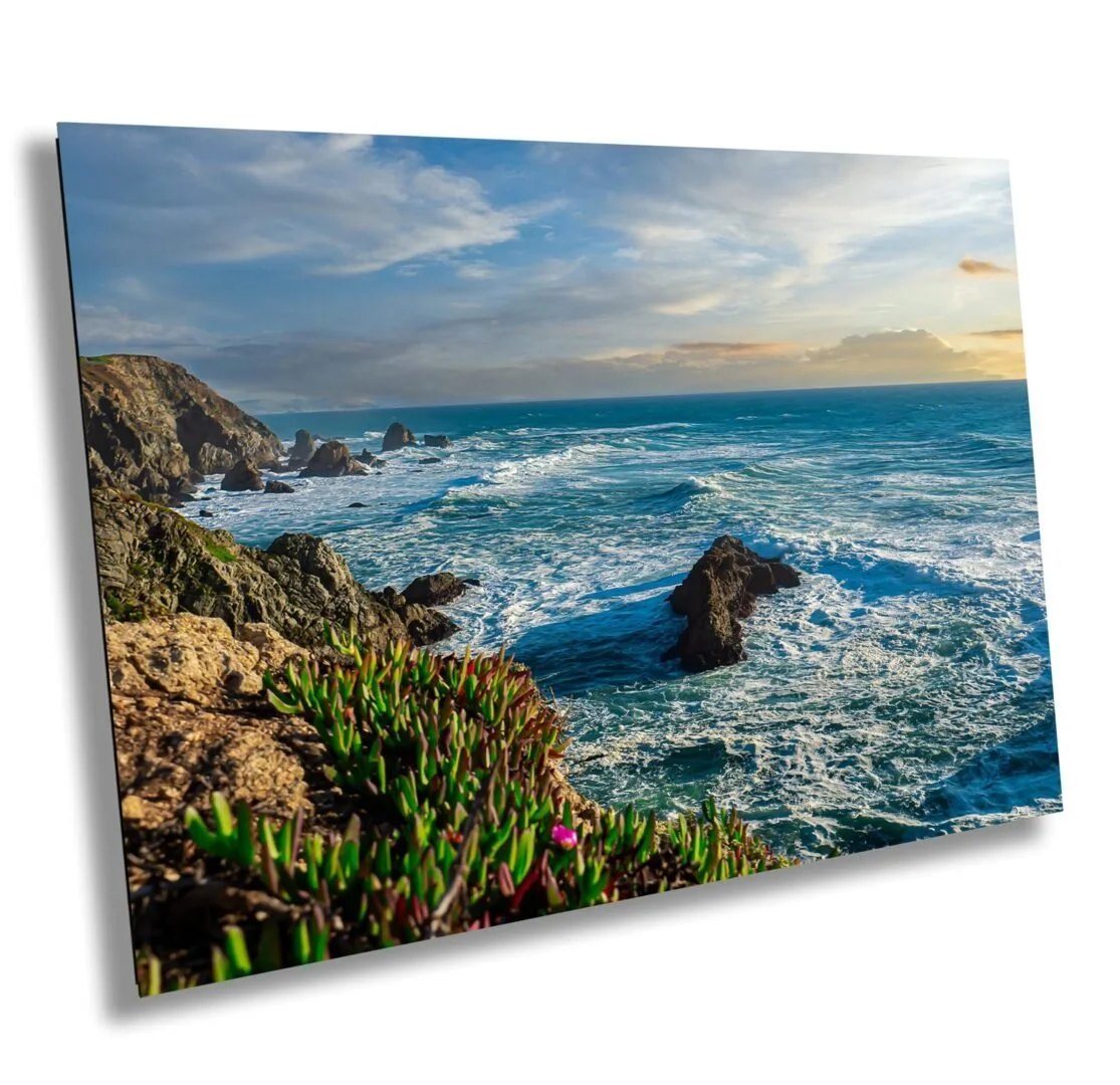 A coastal landscape with rocky cliffs, green plants, and ocean waves under a partly cloudy sky at sunset.