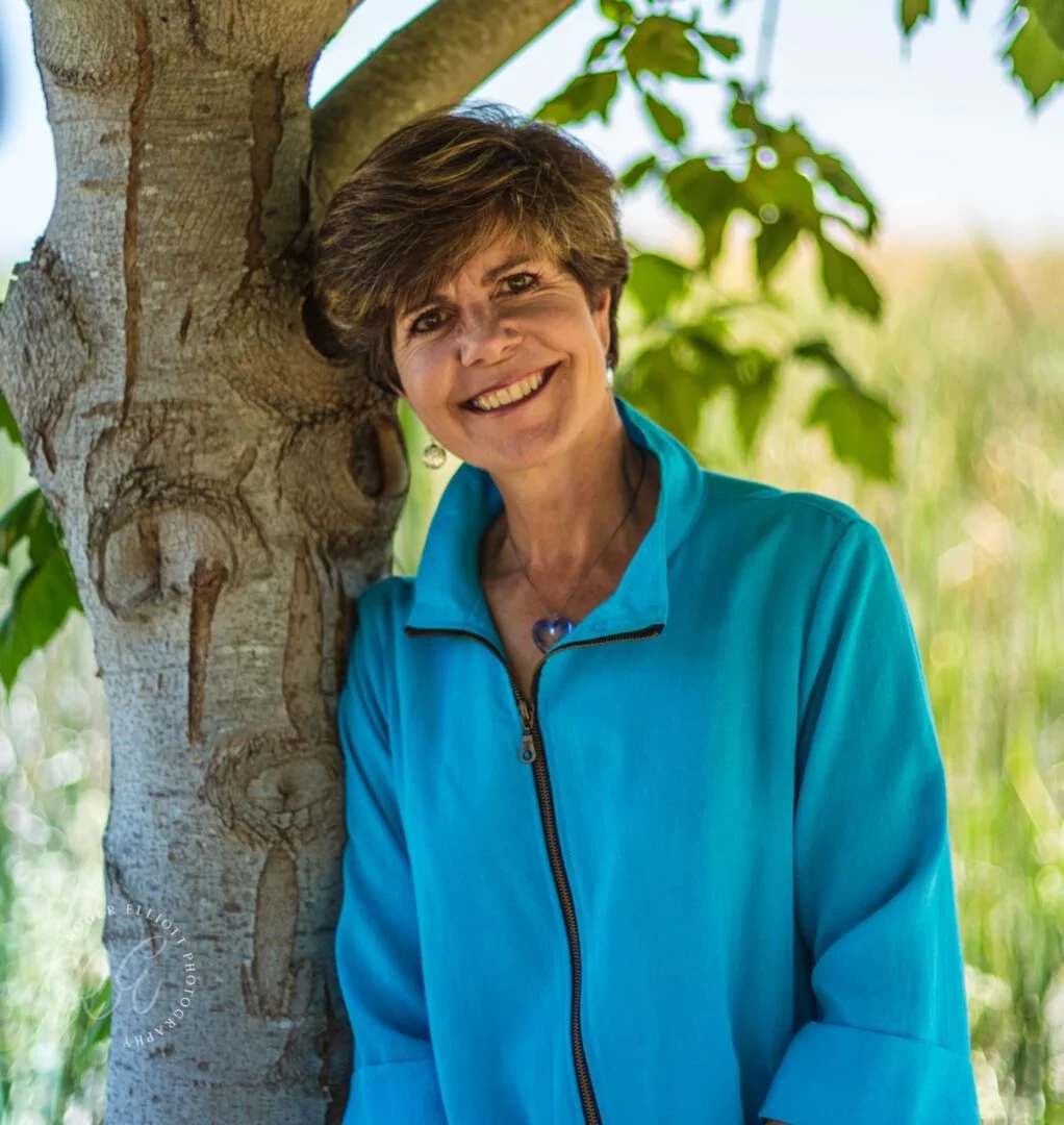 Smiling woman with short brown hair leaning against a tree outdoors, wearing a bright blue jacket, surrounded by green leaves.