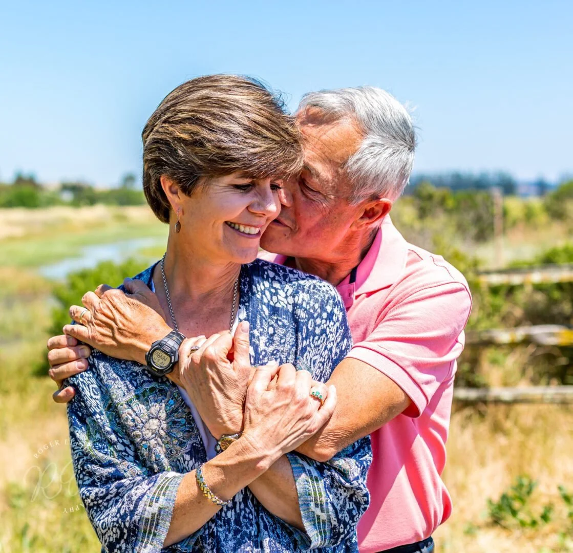 An elderly couple hugging and smiling outdoors on a sunny day with a background of greenery and water.