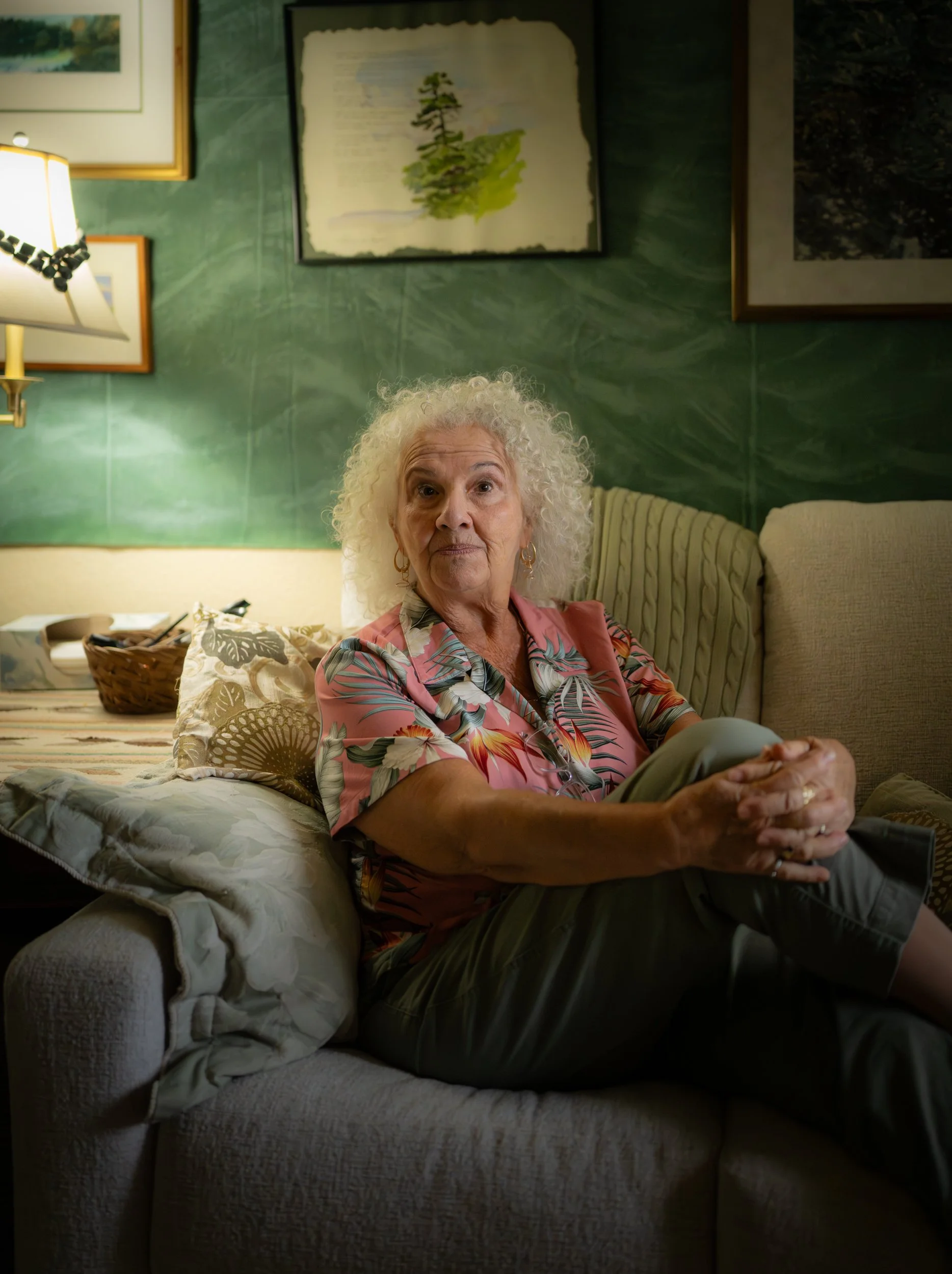 An elderly woman with curly white hair sits on a beige sofa with multiple pillows in a room decorated with green walls and framed pictures. She wears a pink floral blouse and khaki pants, with her hands clasped on her lap, looking slightly to the side.