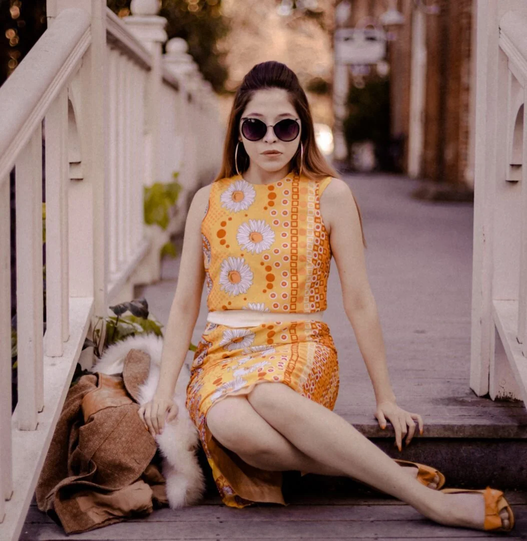 A woman sitting on a wooden bridge, wearing a yellow floral dress and sunglasses, with a small dog resting beside her.