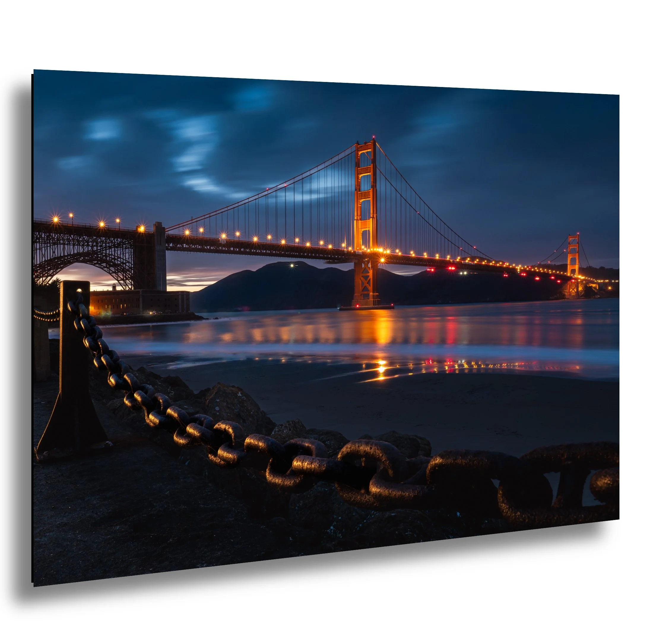 Night view of the Golden Gate Bridge in San Francisco with reflections on the water, illuminated by orange lights, and a cloudy sky overhead.