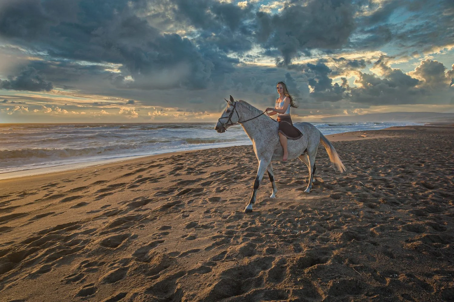 A woman riding a white horse on a sandy beach during sunset, with clouds in the sky and ocean waves in the background.