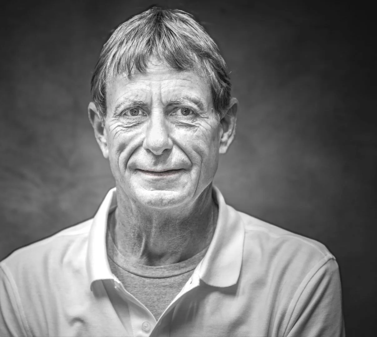 Black and white portrait of an older man with short hair, wearing a collared shirt, smiling gently against a plain dark background.