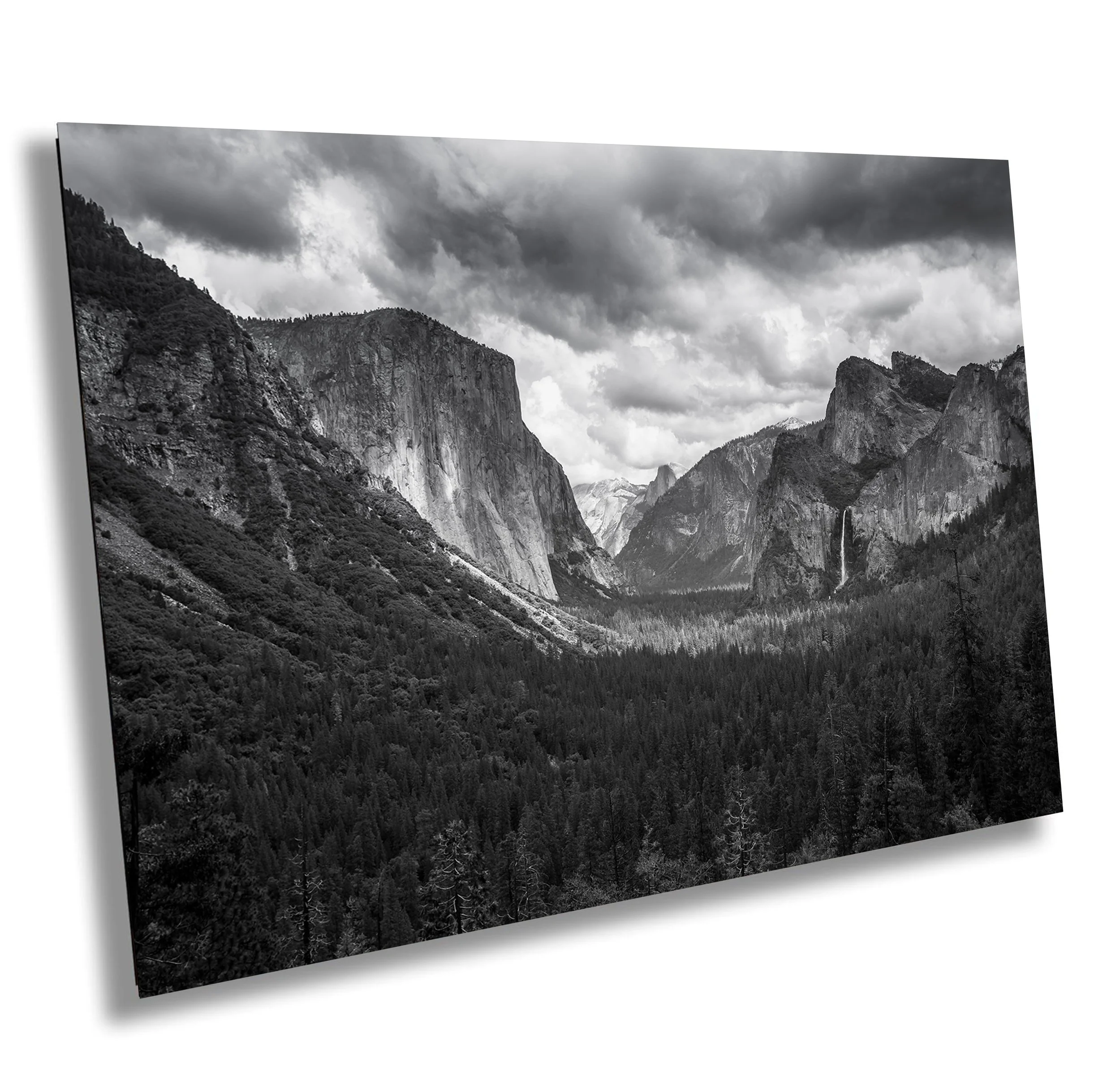 Black and white photograph of a mountainous landscape with tall cliffs, dense forest in the valley, and dramatic cloudy sky.