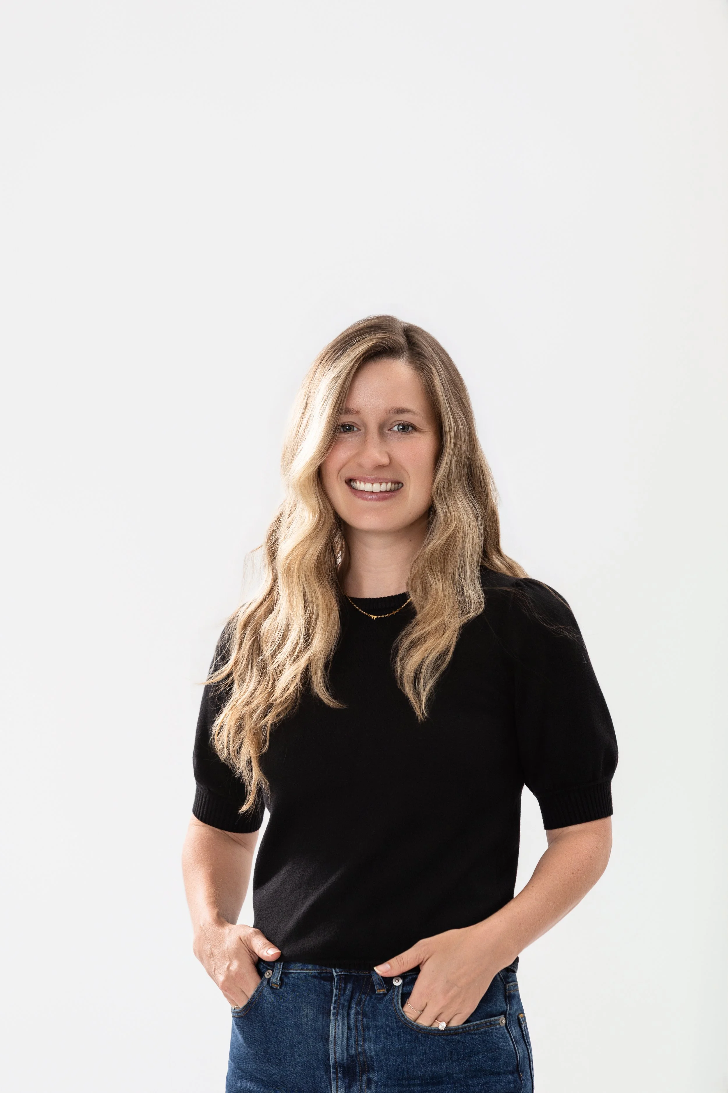 Color portrait of a young woman with wavy hair wearing a a black shirt, smiling, against a white background..