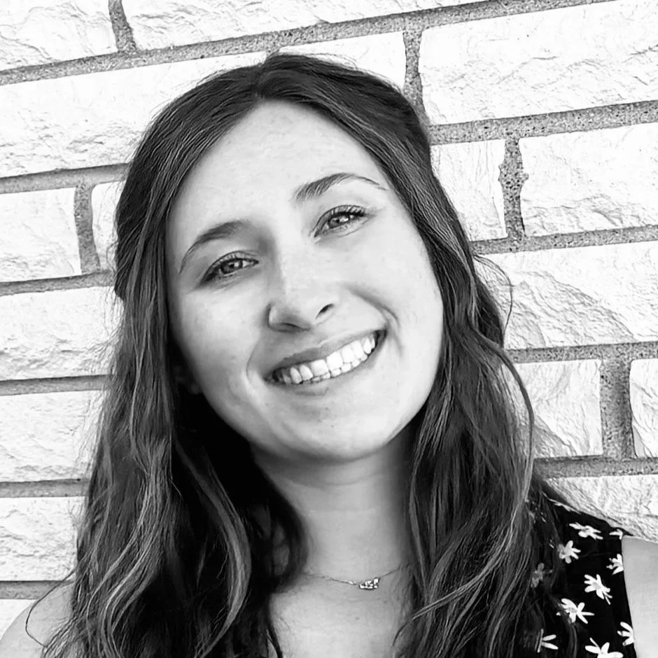 Black and white portrait of a smiling woman with long wavy hair, standing against a brick wall.