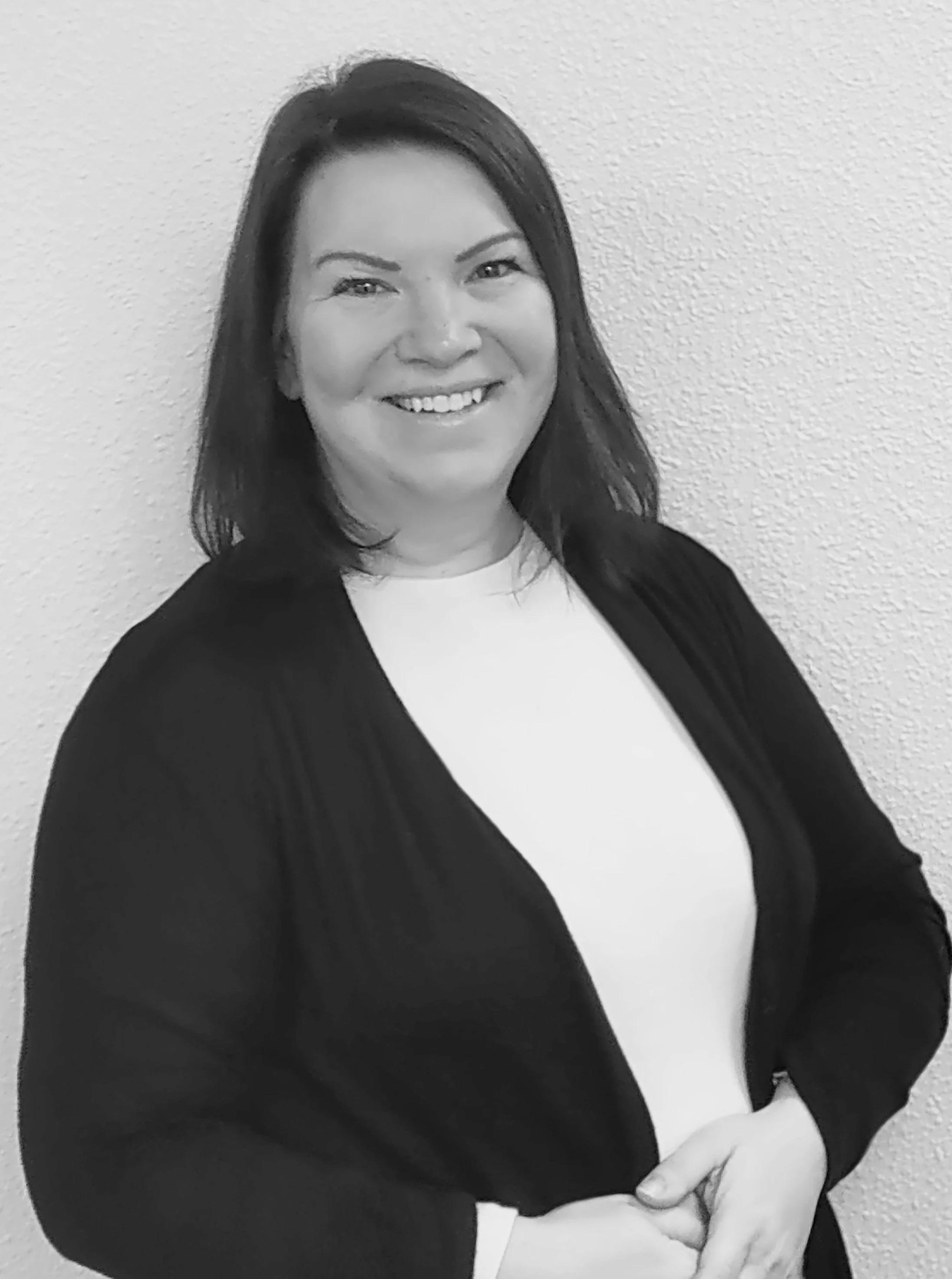 Black and white photo of a woman smiling, with long dark hair, wearing a light-colored top and a dark blazer, standing against a plain wall.