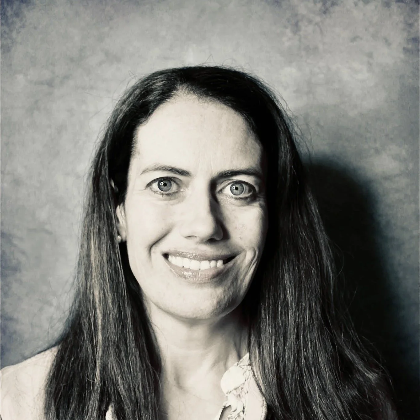 Black and white portrait of a smiling woman with long hair and light-colored eyes, wearing earrings and a floral top.