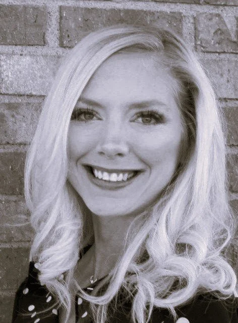 A woman with long, wavy blonde hair smiling in front of a brick wall.