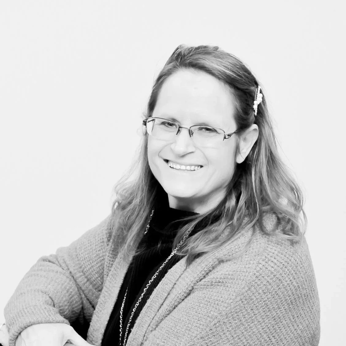 Black and white portrait of a woman with glasses, smiling, wearing a sweater and necklace, with her hair partially clipped back.