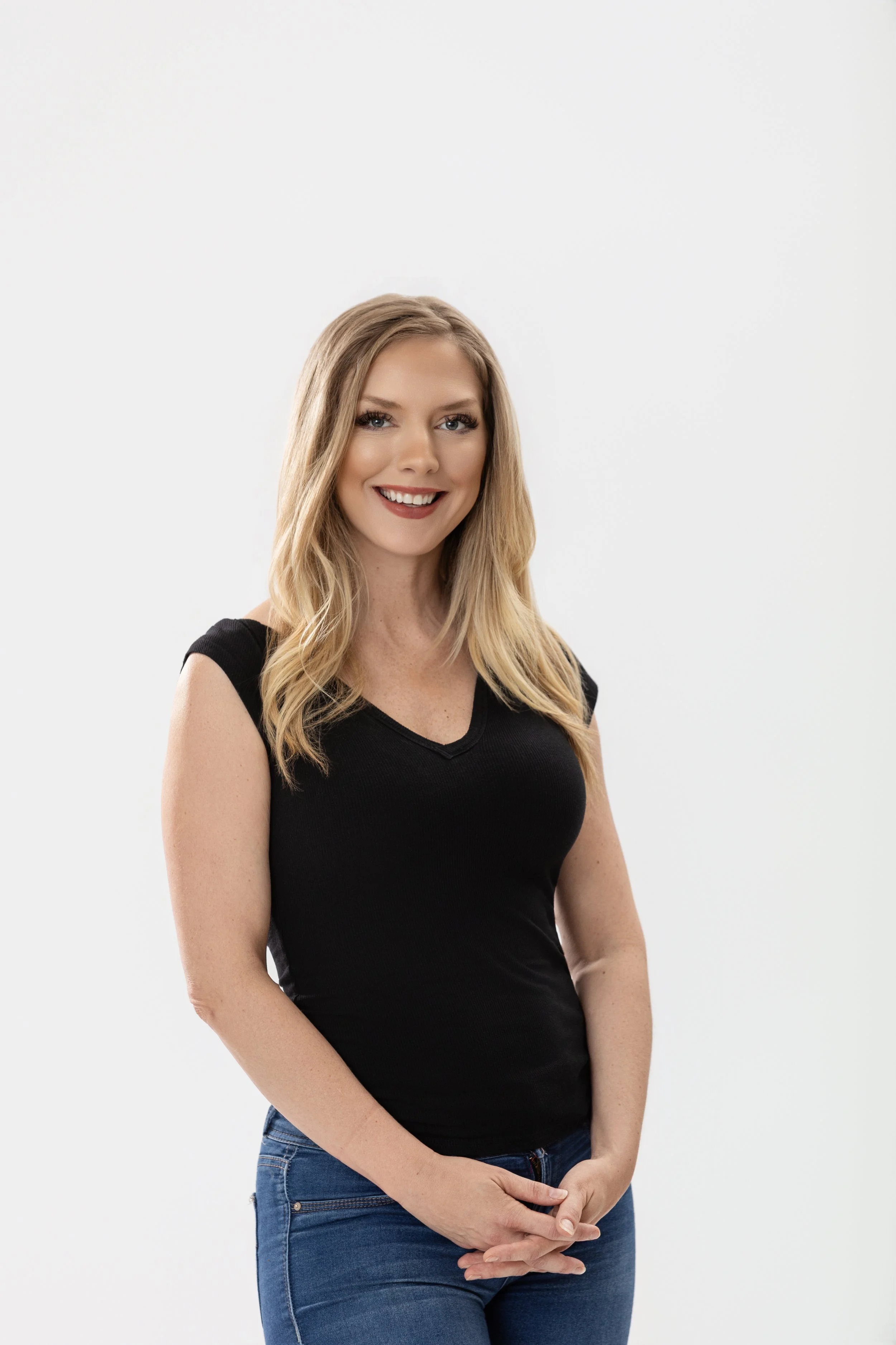 Color portrait of a smiling woman with curled blonde hair, standing and smiling with hands clasped in front of her
