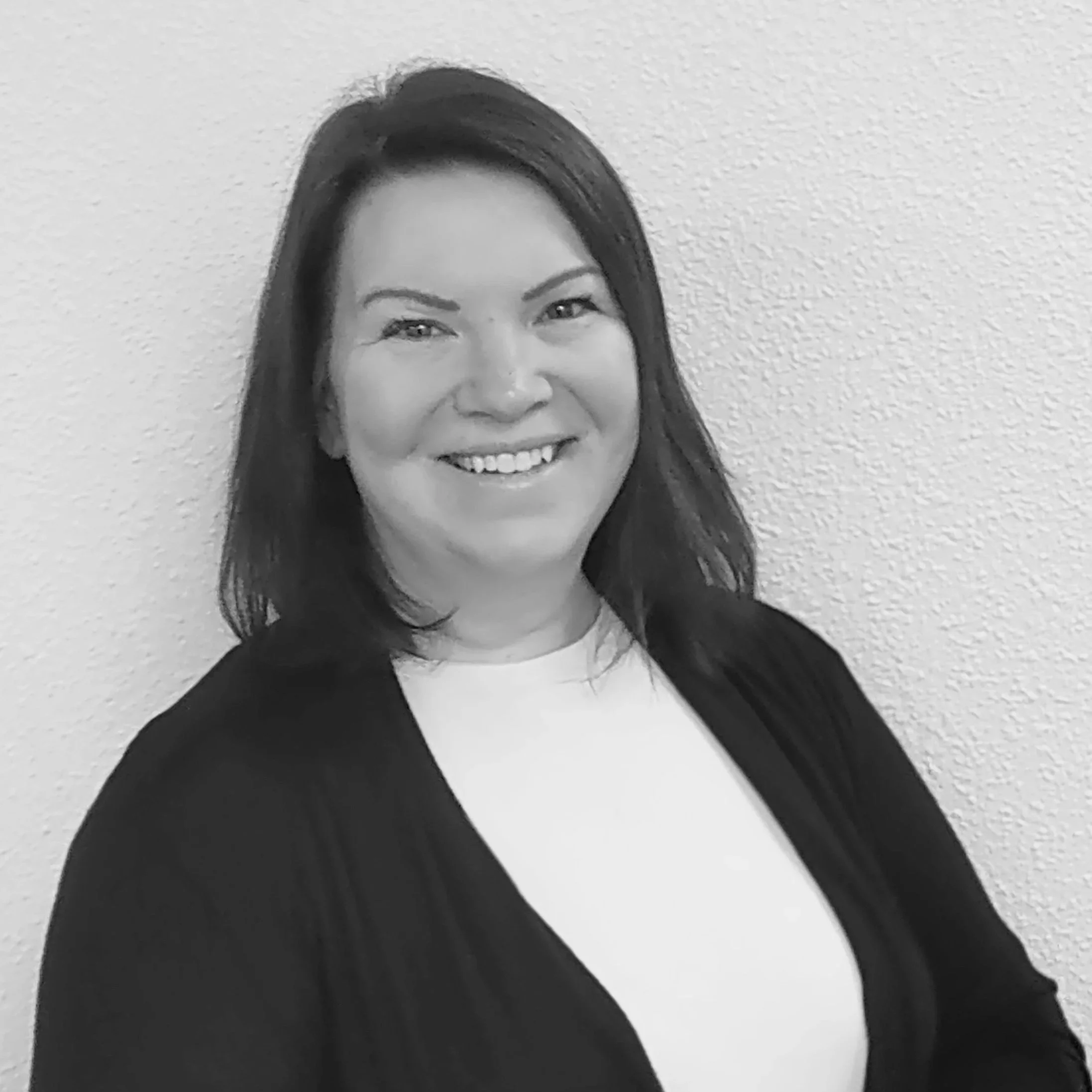 A black and white photo of a woman with long dark hair, smiling, standing against a plain textured wall.