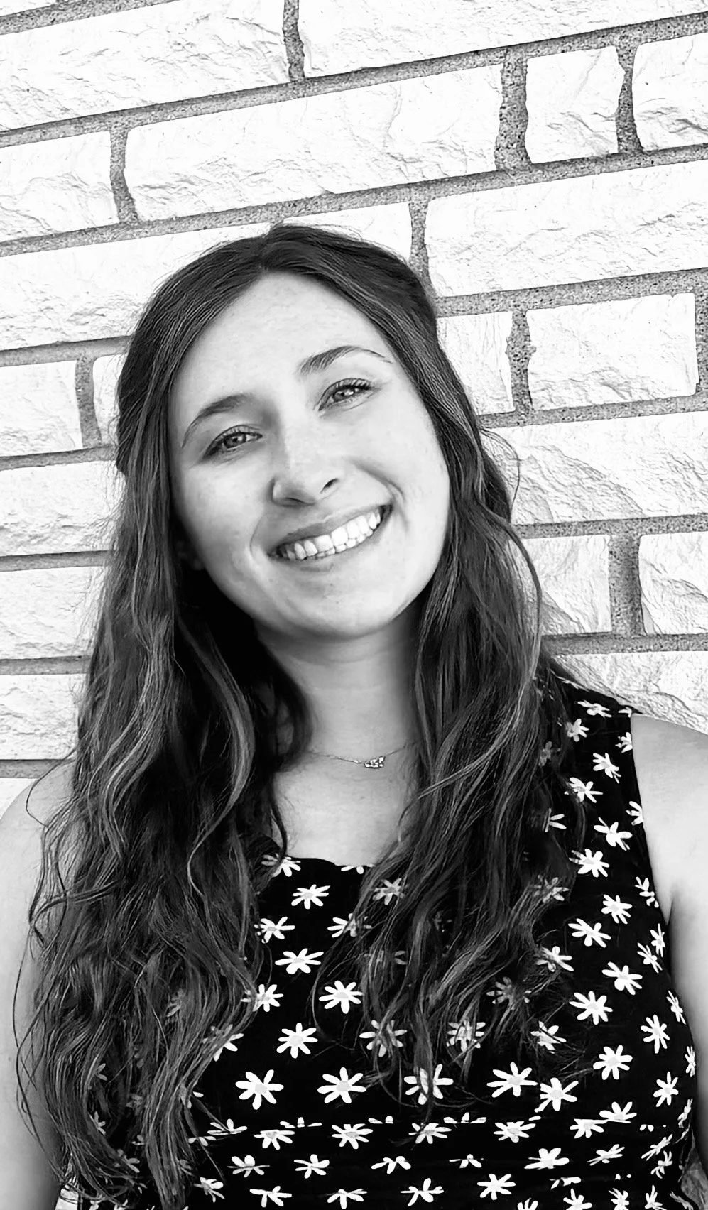 A woman with long wavy hair smiling at the camera, wearing a sleeveless top with a daisy pattern, standing in front of a brick wall.