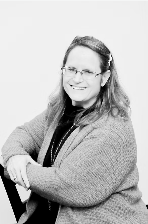 Black and white photo of a woman with glasses, smiling, wearing a cozy sweater and a necklace, sitting with her arms crossed.