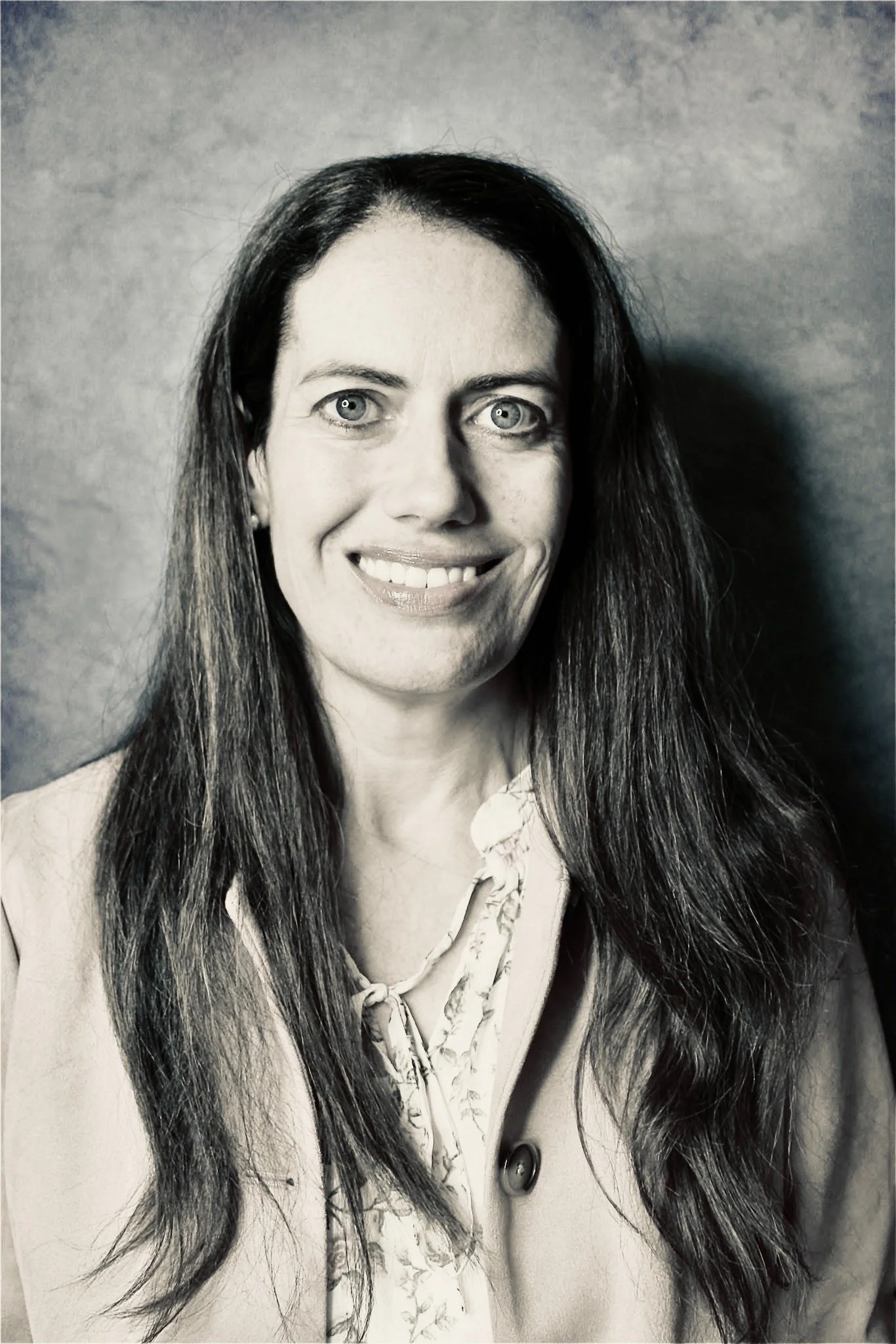 A black and white portrait of a woman with long, wavy hair and light-colored eyes, wearing a light-colored blouse with a tie detail.