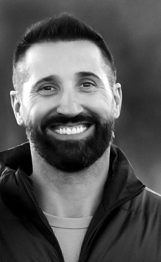 A smiling man with a beard and short hair, wearing a dark jacket, captured in black and white.