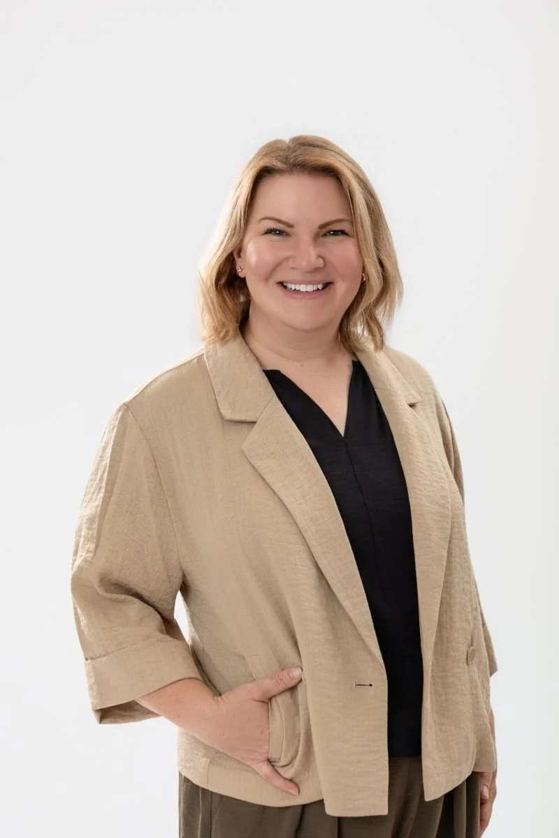 Color photo of a woman smiling, with medium length blonde hair, wearing a light-colored top and a dark blazer, standing against a plain wall.